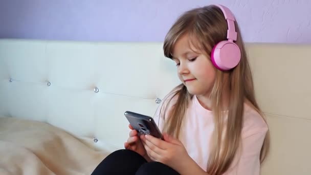 Roztomilé usměvavé děvče ve sluchátkách poslouchá hudbu a trochu tancuje hlavou pomocí chytrého telefonu, sedí doma na posteli, zpomaluje. Vysoce kvalitní 4K záběry - Záběry, video