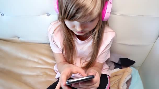 Linda niña preadolescente con auriculares escuchar música y utilizar un teléfono inteligente sentado en una cama en casa, cámara lenta vista superior de cerca clip. Imágenes de alta calidad 4k - Metraje, vídeo