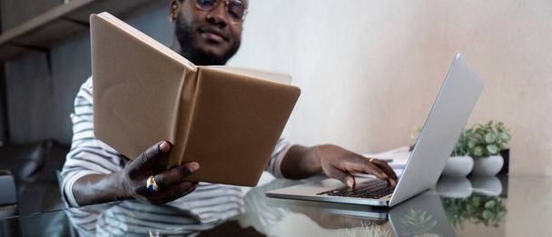 Jonge Afrikaanse man met laptop thuis, zwarte man op zoek naar leesboek ontspannen op de vrije tijd met werk zitten op glazen tafel in de woonkamer. - Foto, afbeelding