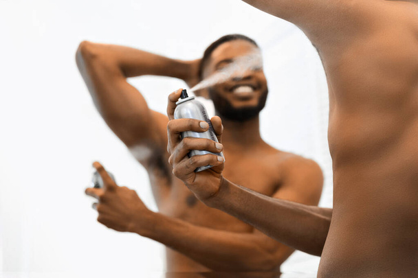 Um afro-americano musculoso sem camisa de fundo branco segura uma garrafa de desodorizante, pulverizando-a sob o braço, mostrando higiene pessoal. - Foto, Imagem