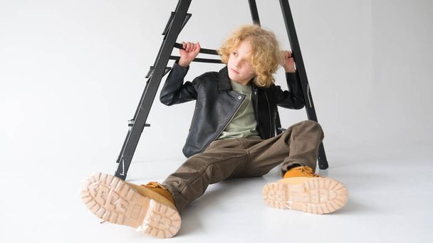 A young boy with voluminous curly hair sits nonchalantly underneath a black ladder, casting a gaze downward. He is wearing a black leather jacket, brown pants, and contrasting yellow boots, creating a relaxed yet stylish look. The ladder suggests an  - Photo, Image