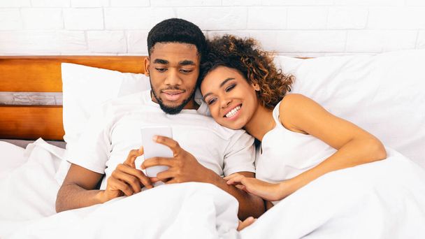 Χαμογελώντας χιλιετή Αφροαμερικανός ζευγάρι χρησιμοποιώντας smartphone μαζί στο κρεβάτι, διαβάζοντας ειδήσεις το πρωί, ελεύθερος χώρος, Πανόραμα - Φωτογραφία, εικόνα