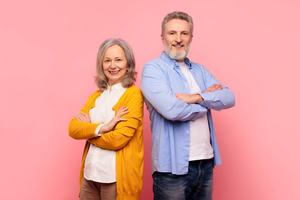 Selbstbewusste Senioren. Reifes Paar posiert mit verschränkten Armen, steht Rücken an Rücken mit Zuversicht, strahlt Glück und Liebe lächelnd in die Kamera, vor rosa Studiohintergrund - Foto, Bild