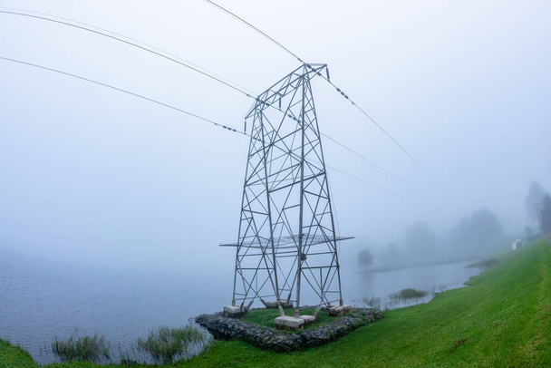Yüksek voltajlı kablosu olan elektrik santralleri kulesi bulutlu bir havada baraj sularının üzerinde su kenarında.. - Fotoğraf, Görsel