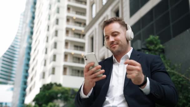 Caucasian businessman listening relaxing music while hold mobile phone. Skilled happy executive manager open music and move to song in lively mood while sitting at green urban city or park. Urbane. - Footage, Video