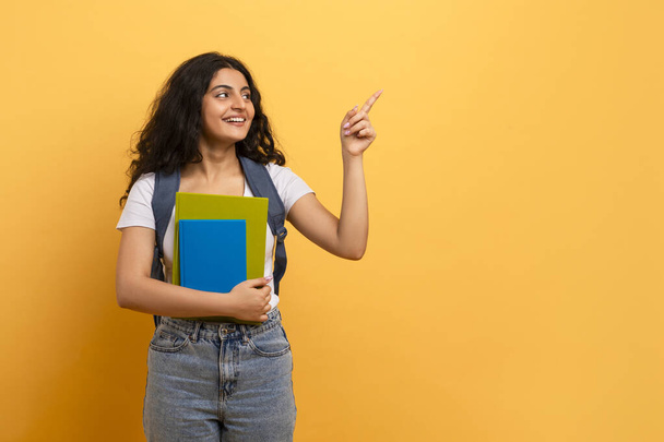 Neugierige Studentin zeigt nach oben, posiert mit blauen Notizbüchern, die Interesse und Ideen signalisieren - Foto, Bild