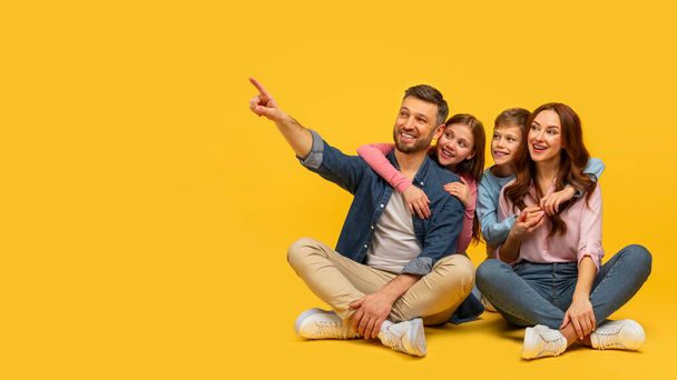 Una familia alegre de cuatro personas sentadas de cerca, el padre señala mientras todo el mundo se ve alegre sobre un fondo amarillo - Foto, Imagen
