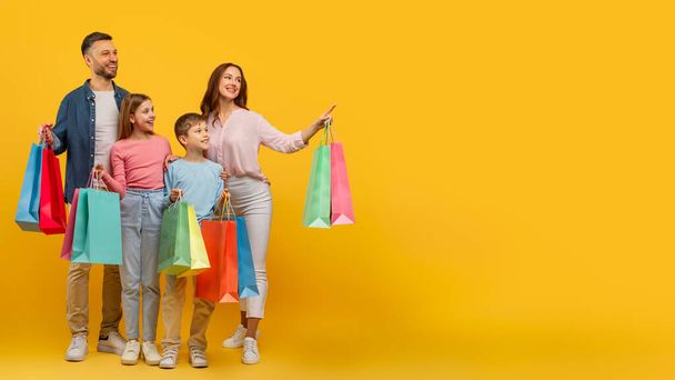 Zabawni rodzice rodziny i dzieci popisują się torbami na zakupy, stojąc na żółtym, wskazując na przestrzeń do kopiowania - Zdjęcie, obraz