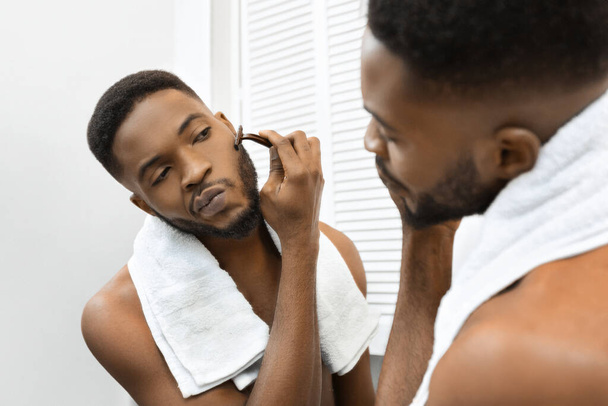 Een jonge Afro-Amerikaanse man met een handdoek om zijn nek kijkt naar zijn spiegelbeeld in de spiegel, scheerbaard - Foto, afbeelding