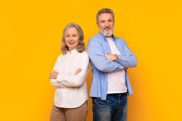 自信を持って引退した夫と妻は,黄色い背景に対して交差した腕と共に立って,黄金時代に自信と友情を示す幸せなシニアカップルのスタジオショット - 写真・画像