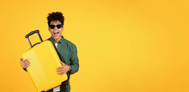 A friendly and joyful african american guy in sunglasses hugs a bright yellow suitcase, displaying happiness and affection for travel on a yellow background - Foto, Imagen