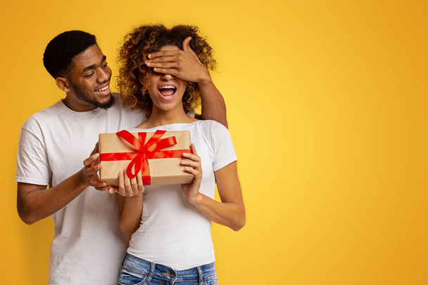 Loving black guy surprising his lovely girlfriend with gift on orange background - Photo, Image