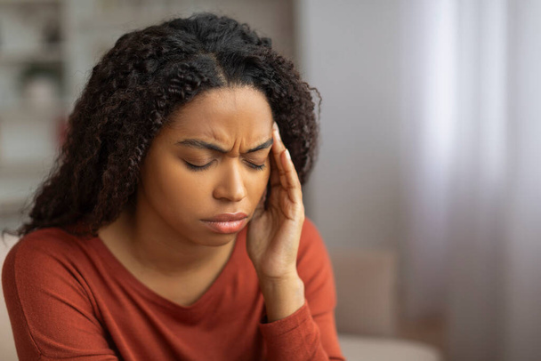 Young black woman in a red top sitting indoors with her eyes closed, pressing her fingertips to her temple in discomfort, african american female suffering headache - Photo, Image