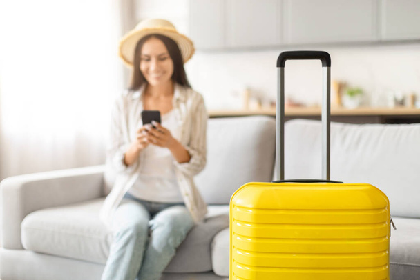 Żółta walizka w centrum uwagi z zamazanym tle, gdzie kobieta korzystająca ze smartfona jest widoczna, rezerwacja taksówki - Zdjęcie, obraz