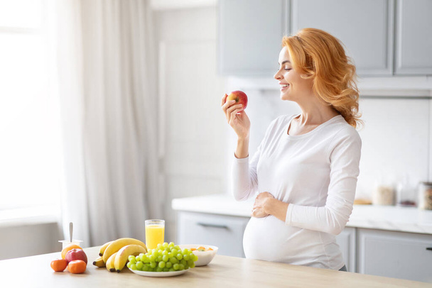 Mujer embarazada saludable en una cocina ordenada seleccionando una fruta entre varias opciones saludables - Foto, imagen