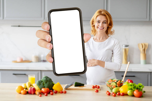 Schwangere blonde Frau hält in einer Küche mit frischen Produkten einen leeren Smartphone-Bildschirm hin - Foto, Bild