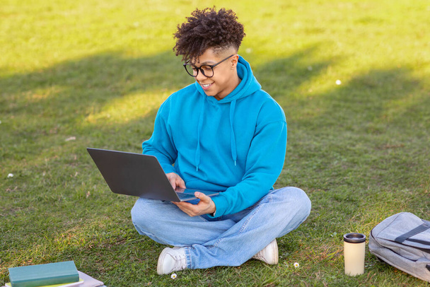 Nenucený mladý africký Američan v jasně modré mikině sedící se zkříženýma nohama na travnatém poli, pracující na laptopu s šálkem kávy poblíž - Fotografie, Obrázek