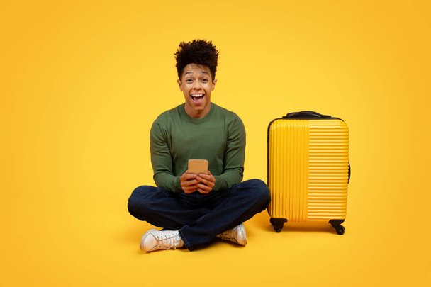 Egy vidám, fiatal afro-amerikai férfi, aki keresztbe tett lábbal ül egy sárga bőrönd mellett, sárga háttérrel, kezében egy okostelefonnal és izgatottan néz. - Fotó, kép