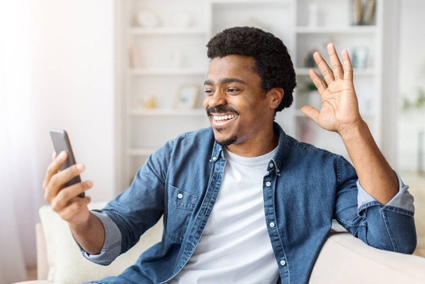 Радостный черный мужчина машет на экран, держа в руках смартфон, участвуя в видеозвонке дома - Фото, изображение