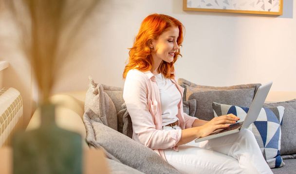 Молодая рыжая улыбающаяся женщина, сидящая на диване и печатающая на ноутбуке. Работа из дома. - Фото, изображение
