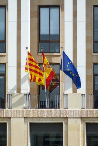 De Spaanse nationale vlag, de Catalaanse Senyera, en de vlag van de Europese Unie wapperen zij aan zij en symboliseren eenheid te midden van diversiteit tegen een moderne architectonische achtergrond. - Foto, afbeelding