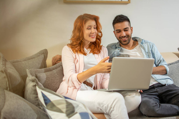 Nettes glückliches Paar benutzt Laptop, während es zu Hause auf dem Sofa sitzt. - Foto, Bild