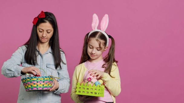 Joyful confident child and mother showing easter baskets on camera, decorating festive arrangements for spring holiday. Happy schoolgirl with bunny ears posing with her mom in studio. Camera A. - Photo, Image