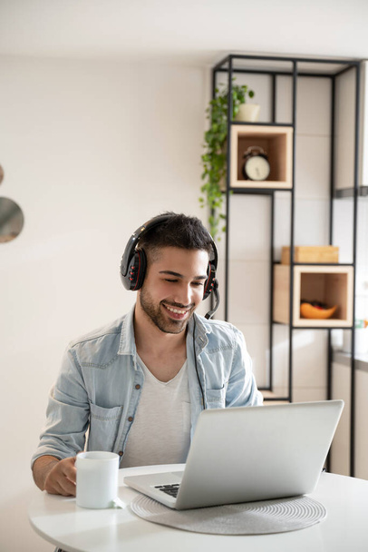 Young happy friendly man working as agent at call center from home while drinking coffee. Wearing headset and using laptop. - Photo, Image