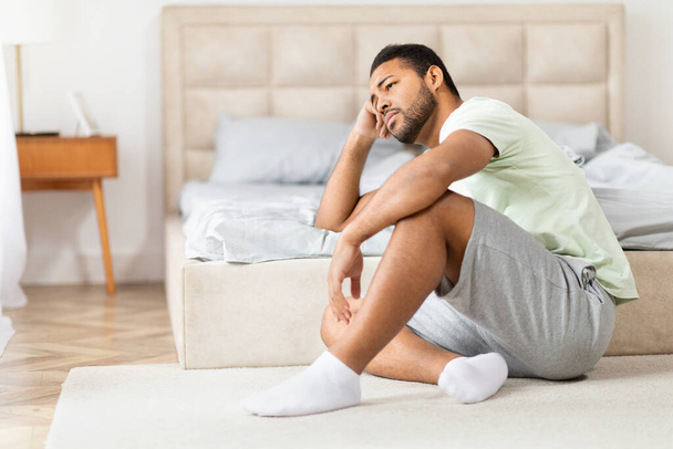 Une image capturant l'homme noir déprimé en tenue décontractée assis jambes croisées sur le tapis à côté d'un lit dans un cadre confortable chambre - Photo, image