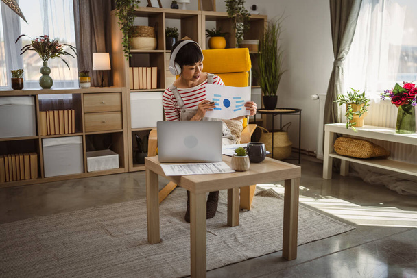 ώριμη γυναίκα Ιαπωνική επιχειρηματίας έχουν βίντεο κλήση χρήση laptop εργασία από το σπίτι online διαβούλευση ή συνέδριο δείχνουν έντυπο έγγραφο προσφορά σύμβασης ή στατιστικά - Φωτογραφία, εικόνα