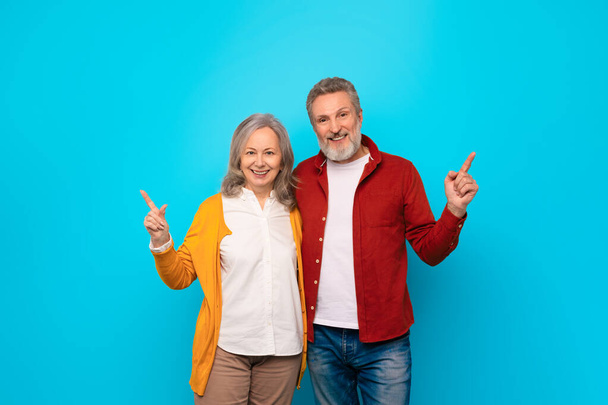 Starsza para przytula się i uśmiecha szczęśliwie, podczas gdy mąż wskazuje palcem w lewo i żona w prawo, wybierając różne strony, reklamując dobrą ofertę na niebieskim tle, studio shot - Zdjęcie, obraz