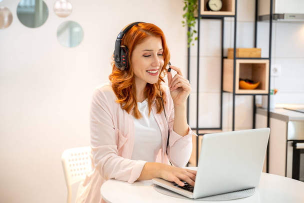 Young happy friendly ginger girl working as agent at call center from home. Wearing headset and typing on laptop. - Photo, Image