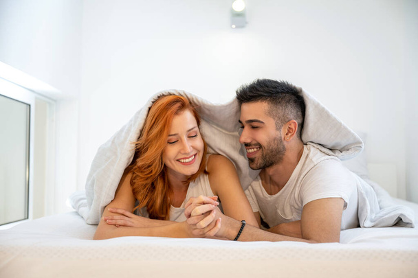 Young smiling couple covered with white blanket, holding hands together and lying in bed at home. Wearing white t-shirt. - Photo, Image