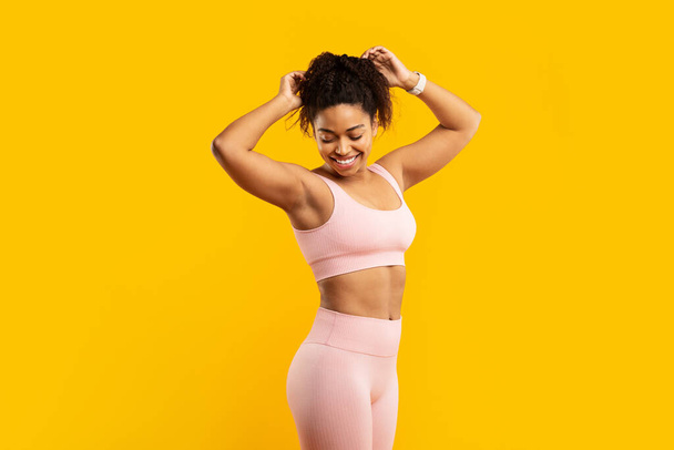 Una vibrante joven afroamericana vestida con un atuendo de entrenamiento rosa prepara felizmente su cabello para una sesión de fitness contra una pared amarilla - Foto, imagen