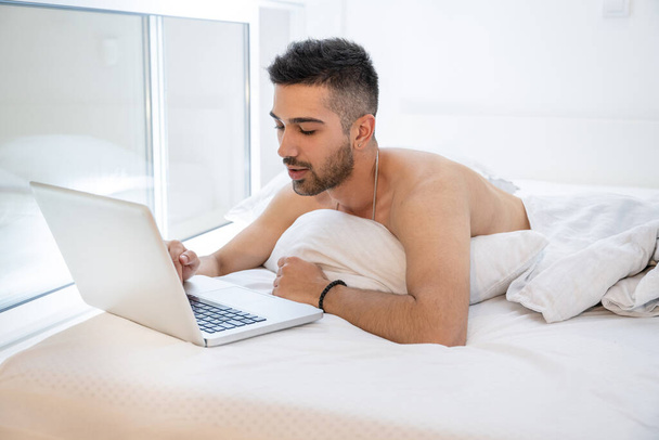 Junger schöner Mann liegt nackt im Bett und surft zu Hause auf Laptop. - Foto, Bild
