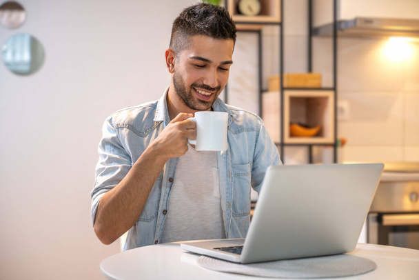 Junger lächelnder Mann, der zu Hause am Laptop arbeitet. Lässige Kleidung tragen und Kaffee trinken. - Foto, Bild