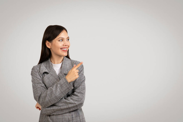 Cheerful businesswoman in classic tweed blazer pointing to her side at free space with toothy smile, suggesting choice or direction, against plain gray background - Photo, Image