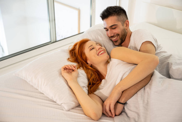 Young smiling hugged couple covered with white blanket, holding hands and lying in bed at home. Wearing white t-shirt. - Photo, Image