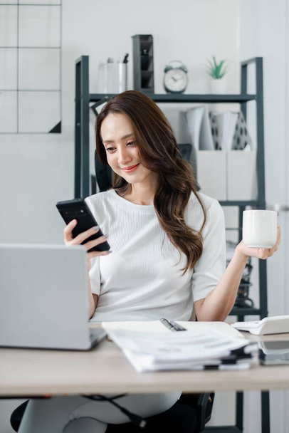 İş kadını elinde bir fincan kahve, önünde bir dizüstü bilgisayar tutarken akıllı telefonunu kontrol eder.. - Fotoğraf, Görsel