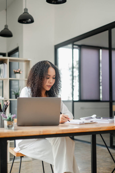 Intensief gefocust werkt een zakenvrouw ijverig op haar laptop aan een ruim bureau in een modern, natuurlijk verlicht kantoor. - Foto, afbeelding