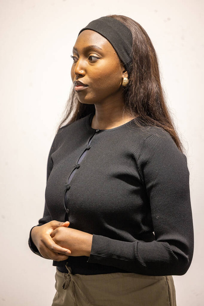 Eine junge Afrikanerin steht vor hellem, neutralem Hintergrund und strahlt Zuversicht und Gelassenheit aus. Sie trägt eine schlichte, aber elegante schwarze Strickjacke und ihr langes Haar ist nach unten gestylt. - Foto, Bild