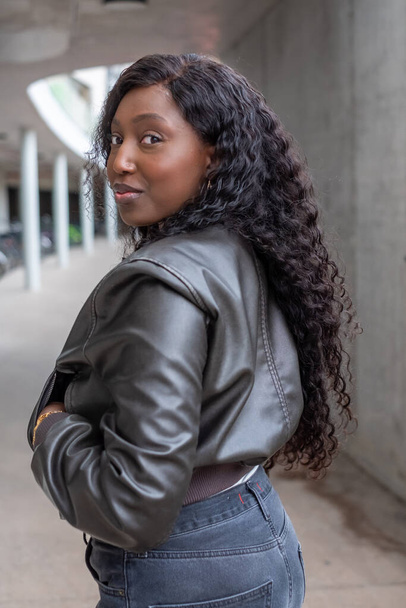 In an urban setting, a confident African woman gives a playful over-the-shoulder look. Her curly hair flows freely, complementing the modern edge of her leather jacket. The soft focus on the concrete - Photo, Image