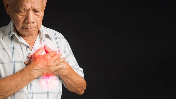 Een oudere Aziatische man in een wit shirt heeft pijn op de linkerborst terwijl hij over een rode achtergrond in de studio staat. Een close-up foto. Ruimte voor tekst. Begrip "ouderen", "geneeskunde" en "gezondheidszorg". - Foto, afbeelding