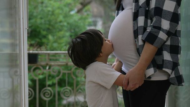 Маленький мальчик выражает любовь, целуя беременный живот матери, нежный момент третьего триместра на домашнем балконе, брат обнимает нерожденного ребенка - Фото, изображение