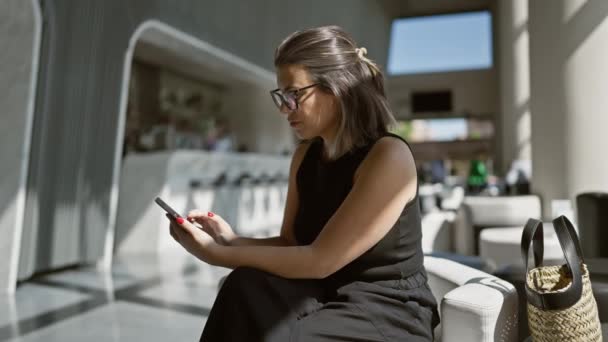 Deliciosa mujer hispana sentada en un moderno hotel, con gafas, usando su teléfono inteligente, sonriendo mientras mira a un lado - Metraje, vídeo