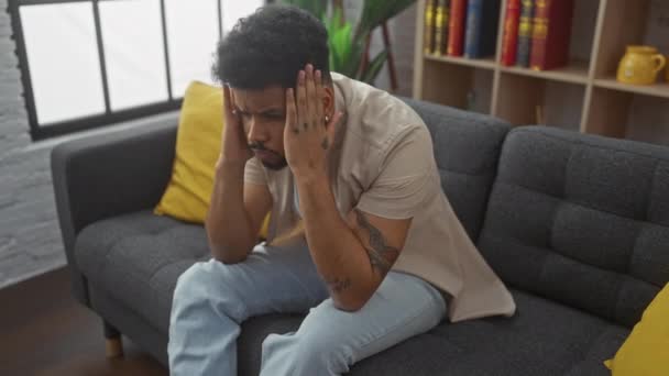 Uomo afroamericano stressato a casa, che soffre di intenso mal di testa sul divano, mano sulla testa in cerca di sollievo da doloroso stress emicrania - Filmati, video