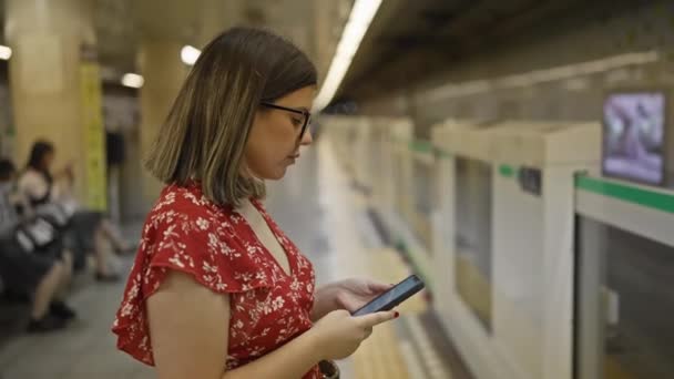 Prachtige Spaanse vrouw, bril op, staat bij het metrostation, anticipeert op haar metro reis, verdiept in haar telefoon - Video