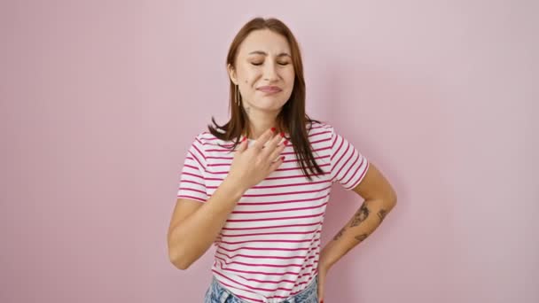 Chica morena joven y dolorida con dolor de garganta, que sufre una infección gripal, agarrando el cuello doloroso sobre un fondo rosa aislado, vestido con una camiseta de rayas - Metraje, vídeo