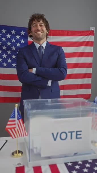 Усміхнений молодий чоловік з схрещеними обіймами стоїть перед американським прапором і голосує виборча скринька в приміщенні. - Кадри, відео
