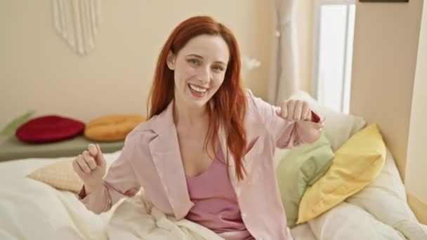 Egy vidám fiatal vörös hajú nő pizsamában élvezi a kényelmes reggelt a jól megvilágított hálószobájában. - Felvétel, videó